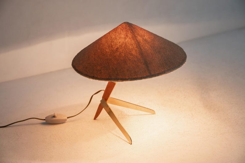 May include: A mid-century table lamp with a conical, brown lampshade. The lamp has a wooden base with three legs and a red support. The lamp is turned on, and the cord has a switch.