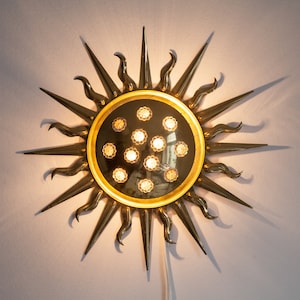 以下が含まれることがあります： 中央に丸い鏡が付いた、金色をした太陽の形をした壁掛け照明。鏡は金色の枠で囲まれており、12個の小さな透明なガラス電球で囲まれています。