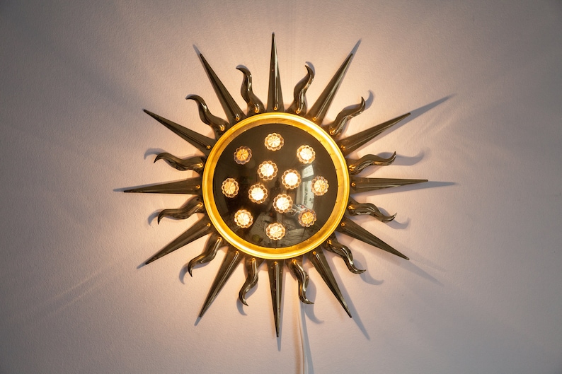以下が含まれることがあります： 中央に丸い鏡が付いた、金色をした太陽の形をした壁掛け照明。鏡は金色の枠で囲まれており、12個の小さな透明なガラス電球で囲まれています。