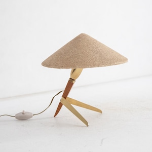 May include: A mid-century modern table lamp with a conical beige shade. The lamp has a wooden and brass base with three legs. The lamp has a brown cord and a small, round on/off switch. The lamp is a decorative lighting fixture.