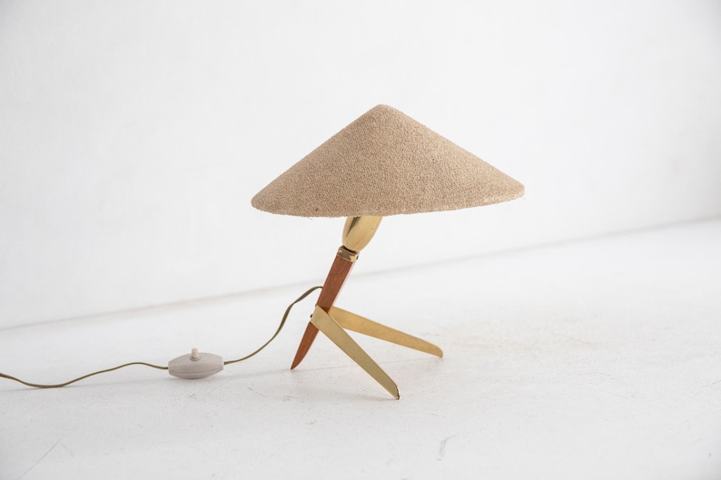 May include: A mid-century modern table lamp with a conical beige shade. The lamp has a wooden and brass base with three legs. The lamp has a brown cord and a small, round on/off switch. The lamp is a decorative lighting fixture.