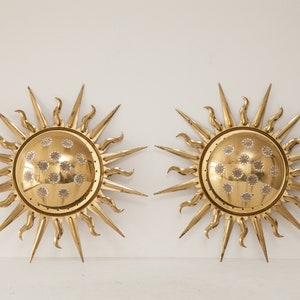 以下が含まれることがあります： 装飾的なテクスチャのある円形のセンターを持つ、ゴールドのサンバーストウォール sconces のペア。各 sconces は、16 本のポイントを持つスターバーストデザインです。