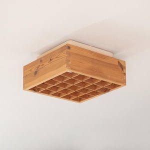 Puede incluir: Una lámpara de techo cuadrada de madera. La lámpara está hecha de madera de color claro y presenta un patrón de rejilla en la parte inferior. Está montada en un techo blanco.