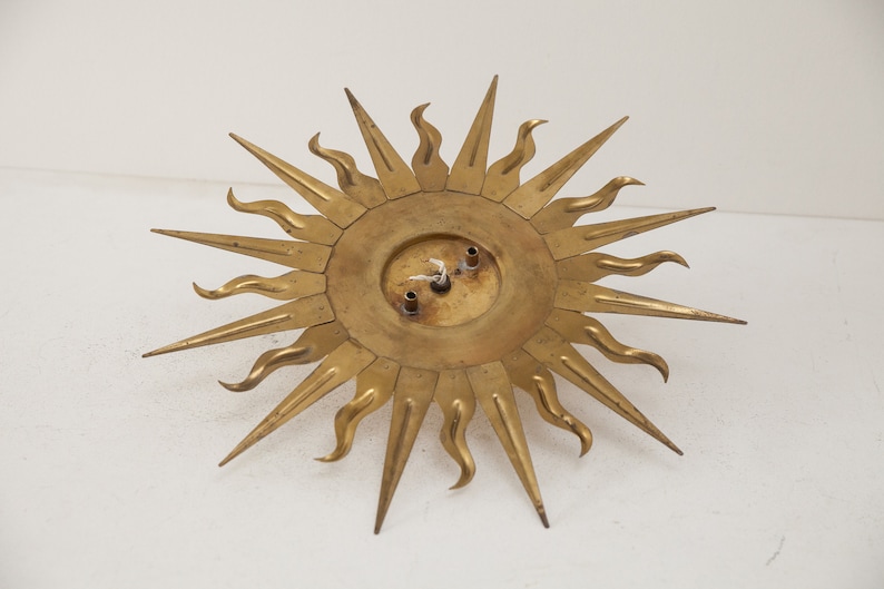 以下が含まれることがあります： 円形のセンターと尖った光線を持つ、金色の金属製のサンバーストウォールデコレーション。光線は波状のデザインになっています。