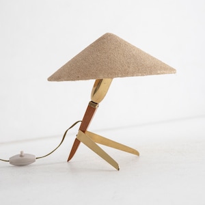 May include: A mid-century table lamp with a triangular beige fabric shade. The lamp has a brass and wood base with three legs. The lamp has a brown cord and a round, light-coloured switch.