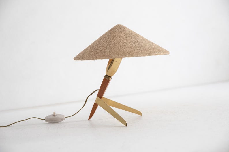 May include: A mid-century table lamp with a triangular beige fabric shade. The lamp has a brass and wood base with three legs. The lamp has a brown cord and a round, light-coloured switch.