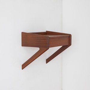 May include: A mid-century modern wall-mounted bedside table. The table is made of dark brown wood and features a drawer and a shelf. The legs are angled, giving it a unique design. It is mounted on a white wall.