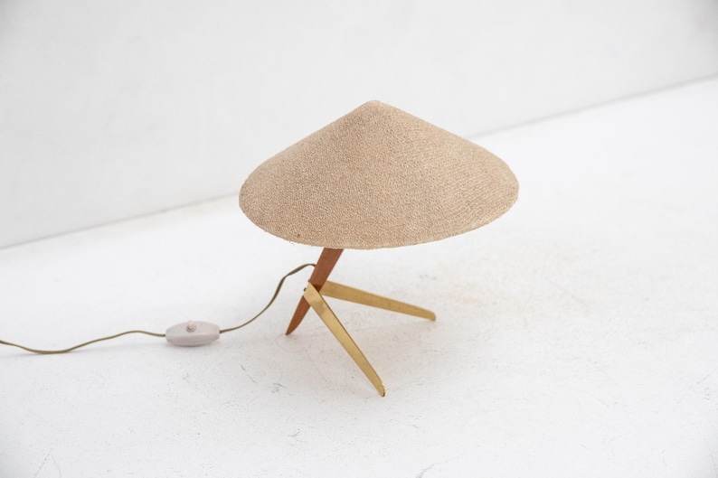 May include: A mid-century modern table lamp with a beige, conical shade. The lamp has a wooden stem and brass legs. The lamp has a cord with an on/off switch. The lamp is a decorative lighting fixture.