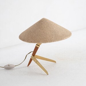 May include: A mid-century table lamp with a conical beige shade. The lamp has a wooden and brass tripod base, a brown wooden stem, and a beige cord with a grey switch. The lamp is designed for indoor use.