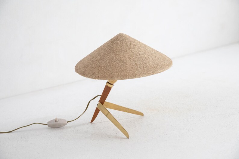 May include: A mid-century table lamp with a conical beige shade. The lamp has a wooden and brass tripod base, a brown wooden stem, and a beige cord with a grey switch. The lamp is designed for indoor use.