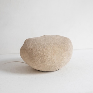Puede incluir: Una lámpara de mesa con una forma irregular, redondeada y similar a una piedra, con un exterior texturizado de color beige arena. La lámpara cuenta con un cable eléctrico fino y claro que sale de su base, apoyada sobre una superficie neutra.