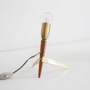 May include: A mid-century modern table lamp with a clear lightbulb, brass accents, and a wooden base. The lamp has a three-legged base and a cord with an on/off switch. The lamp is approximately 25 cm tall.