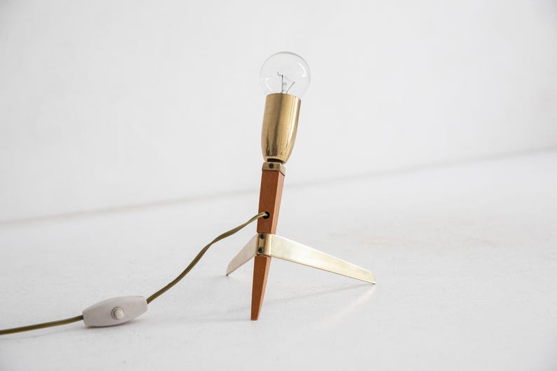 May include: A mid-century modern table lamp with a clear lightbulb, brass accents, and a wooden base. The lamp has a three-legged base and a cord with an on/off switch. The lamp is approximately 25 cm tall.