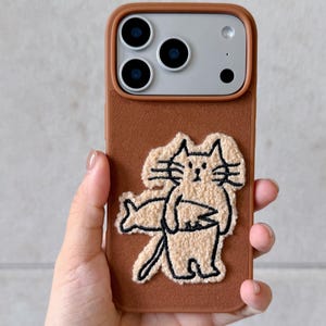Puede incluir: Funda de tel&eacute;fono marr&oacute;n con un dise&ntilde;o de gato bordado en beige. La funda tiene un borde marr&oacute;n y una ilustraci&oacute;n de gato beige. La marca "SookieOfficial" est&aacute; impresa en la parte superior. La funda del tel&eacute;fono la sostiene una persona.