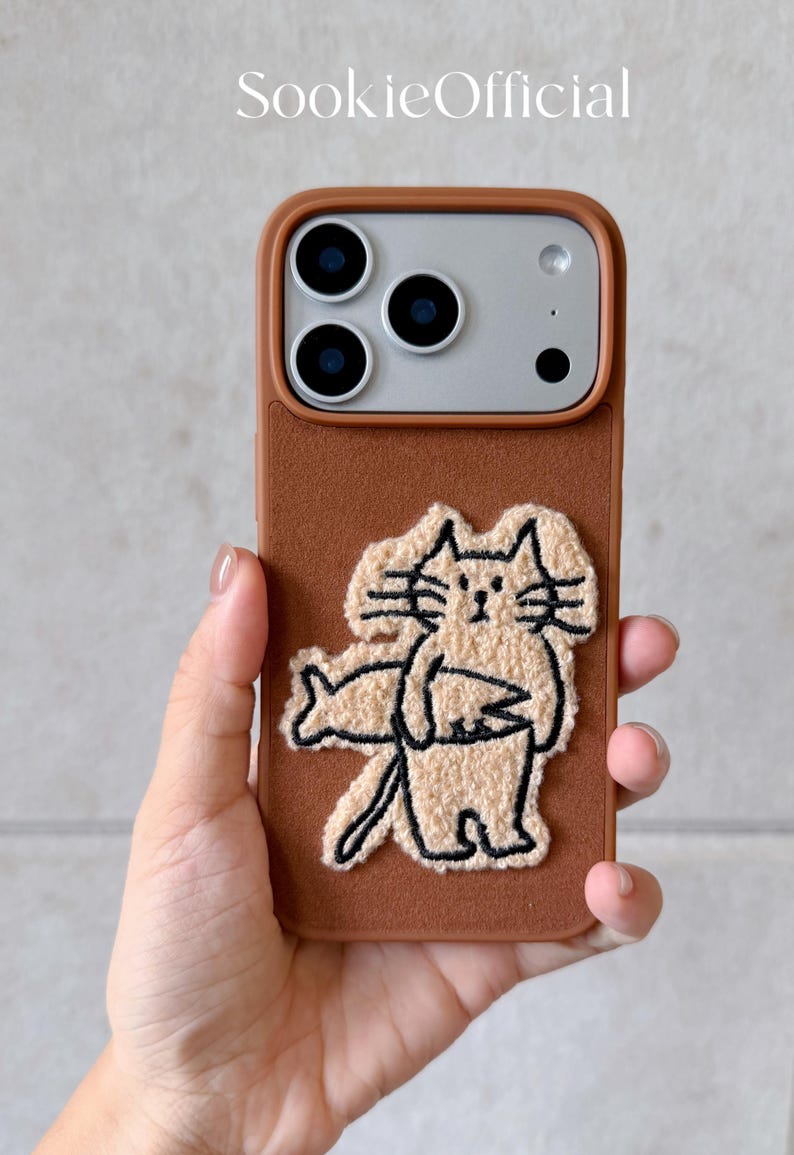 Puede incluir: Funda de tel&eacute;fono marr&oacute;n con un dise&ntilde;o de gato bordado en beige. La funda tiene un borde marr&oacute;n y una ilustraci&oacute;n de gato beige. La marca "SookieOfficial" est&aacute; impresa en la parte superior. La funda del tel&eacute;fono la sostiene una persona.