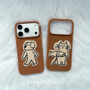 Puede incluir: Dos fundas marrones para tel&eacute;fono con dise&ntilde;os de animales bordados. Una funda presenta un perro, la otra un gato. Las fundas tienen un borde marr&oacute;n y est&aacute;n sobre un fondo blanco y esponjoso. El texto "SookieOfficial" est&aacute; en la parte inferior.
