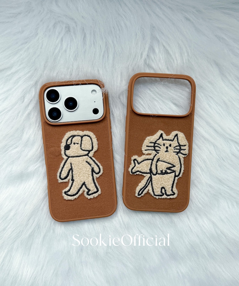 Puede incluir: Dos fundas marrones para tel&eacute;fono con dise&ntilde;os de animales bordados. Una funda presenta un perro, la otra un gato. Las fundas tienen un borde marr&oacute;n y est&aacute;n sobre un fondo blanco y esponjoso. El texto "SookieOfficial" est&aacute; en la parte inferior.