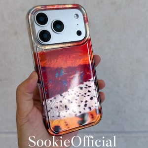 May include: A clear phone case with a vibrant abstract design in shades of red, orange, and black. The case has a silver camera module and the text "SookieOfficial" at the bottom. The case is designed to fit a smartphone.