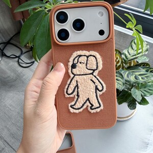 Puede incluir: Una funda de tel&eacute;fono marr&oacute;n con un aplique de perro texturizado marr&oacute;n claro. La funda tiene un borde marr&oacute;n claro y un recorte para la c&aacute;mara. La funda del tel&eacute;fono se muestra frente a plantas verdes.