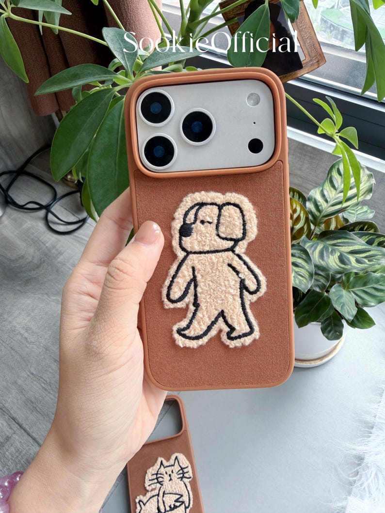 Puede incluir: Una funda de tel&eacute;fono marr&oacute;n con un aplique de perro texturizado marr&oacute;n claro. La funda tiene un borde marr&oacute;n claro y un recorte para la c&aacute;mara. La funda del tel&eacute;fono se muestra frente a plantas verdes.