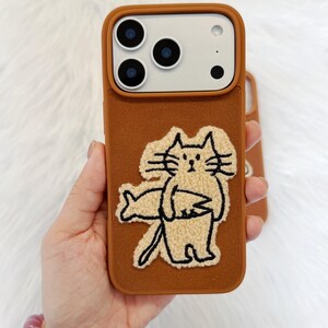 Puede incluir: Funda de tel&eacute;fono marr&oacute;n con un acabado texturizado y un dise&ntilde;o de gato de dibujos animados. El gato es beige con contornos negros y sostiene un pez. La funda tiene un recorte para la c&aacute;mara y el texto "SookieOfficial".