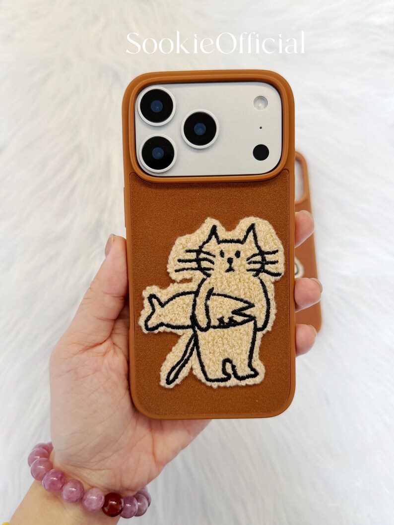 Puede incluir: Funda de tel&eacute;fono marr&oacute;n con un acabado texturizado y un dise&ntilde;o de gato de dibujos animados. El gato es beige con contornos negros y sostiene un pez. La funda tiene un recorte para la c&aacute;mara y el texto "SookieOfficial".