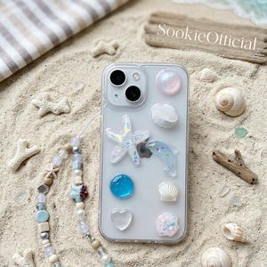 May include: Clear phone case decorated with a starfish, dolphin, and other ocean-themed embellishments. A beaded phone charm and seashells are scattered on the sand. The text "SookieOfficial" is visible on a wooden plank.
