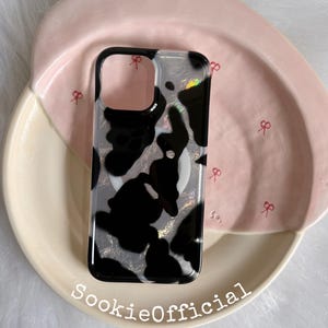 May include: A rectangular phone case featuring a black and white cow print pattern with a subtle iridescent shimmer. The case has a black frame and a circular magnetic ring on the back. The text SookieOfficial is printed in white at the bottom.