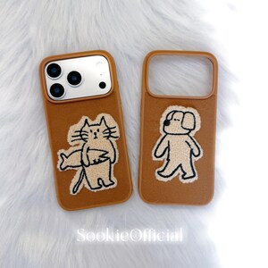 Puede incluir: Dos fundas marrones para tel&eacute;fono con dise&ntilde;os bordados. Una funda presenta un gato sosteniendo un pez, y la otra un perro. Las fundas tienen un aspecto suave y texturizado, sobre un fondo blanco y esponjoso. El texto "SookieOfficial" es visible.