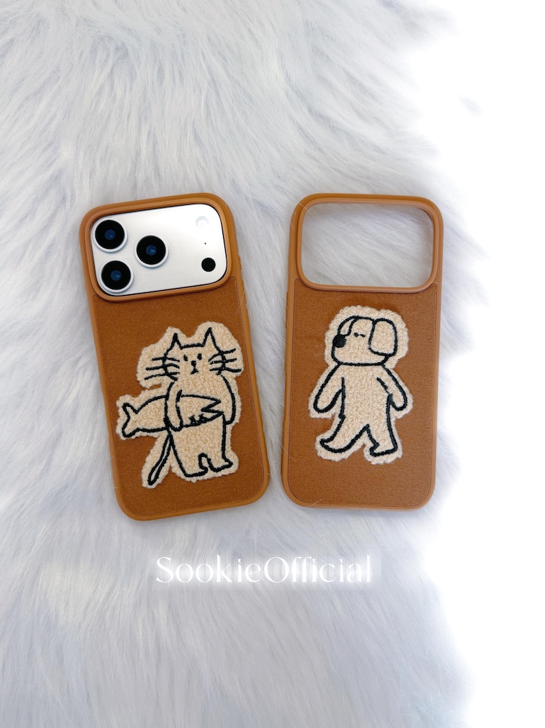 Puede incluir: Dos fundas marrones para tel&eacute;fono con dise&ntilde;os bordados. Una funda presenta un gato sosteniendo un pez, y la otra un perro. Las fundas tienen un aspecto suave y texturizado, sobre un fondo blanco y esponjoso. El texto "SookieOfficial" es visible.