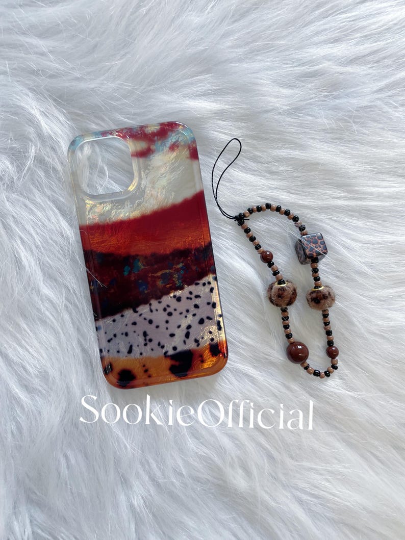 May include: A clear phone case with a layered design in shades of red, orange, and white, with a black beaded wrist strap. The case has the text "SookieOfficial" at the bottom.