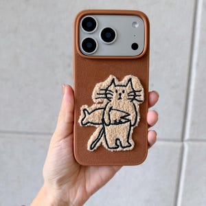 Puede incluir: Funda de tel&eacute;fono marr&oacute;n con dise&ntilde;o de gato. La funda tiene una superficie texturizada y presenta una ilustraci&oacute;n de gato beige. El texto "SookieOfficial" est&aacute; en la parte superior. La funda est&aacute; dise&ntilde;ada para un tel&eacute;fono inteligente.