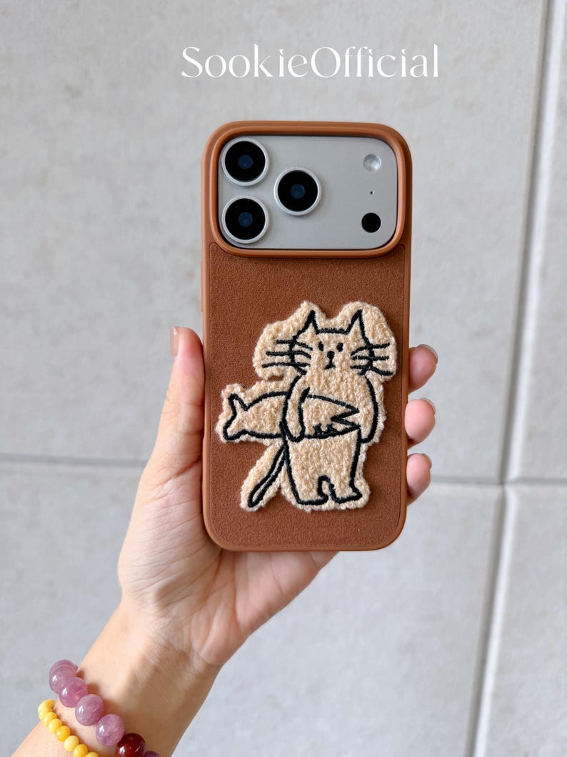Puede incluir: Funda de tel&eacute;fono marr&oacute;n con dise&ntilde;o de gato. La funda tiene una superficie texturizada y presenta una ilustraci&oacute;n de gato beige. El texto "SookieOfficial" est&aacute; en la parte superior. La funda est&aacute; dise&ntilde;ada para un tel&eacute;fono inteligente.