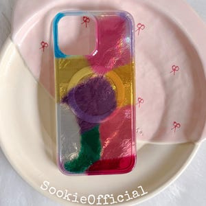 Peut inclure: Une coque de téléphone rectangulaire présentant un motif abstrait multicolore avec des taches de bleu, jaune, violet, vert et rose. La coque comprend un anneau magnétique circulaire au centre et une découpe carrée pour l'appareil photo. Le texte SookieOfficial est visible.