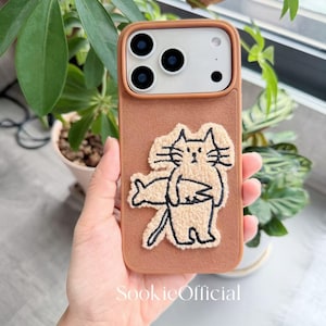 Puede incluir: Funda de tel&eacute;fono marr&oacute;n con un dise&ntilde;o de gato crema sosteniendo un pez. La funda tiene una textura en relieve y un recorte para la c&aacute;mara. La marca "SookieOfficial" est&aacute; impresa en la parte inferior.