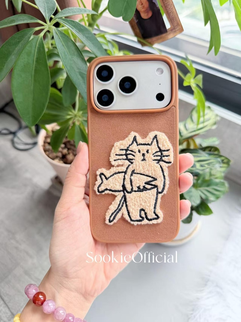 Puede incluir: Funda de tel&eacute;fono marr&oacute;n con un dise&ntilde;o de gato crema sosteniendo un pez. La funda tiene una textura en relieve y un recorte para la c&aacute;mara. La marca "SookieOfficial" est&aacute; impresa en la parte inferior.