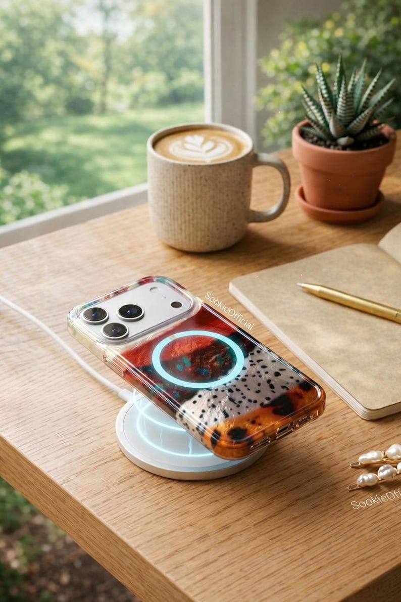 May include: A clear phone case with a colorful design, resting on a wireless charger. The case features a pattern of red, orange, and white. A mug of coffee and a small potted plant are also on the wooden surface.
