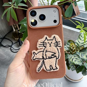 Puede incluir: Una funda de tel&eacute;fono marr&oacute;n con un dise&ntilde;o de gato beige texturizado. La funda tiene un recorte para la c&aacute;mara y el texto "SookieOfficial" en la parte inferior. La funda del tel&eacute;fono se sostiene en una mano, con plantas verdes en el fondo.