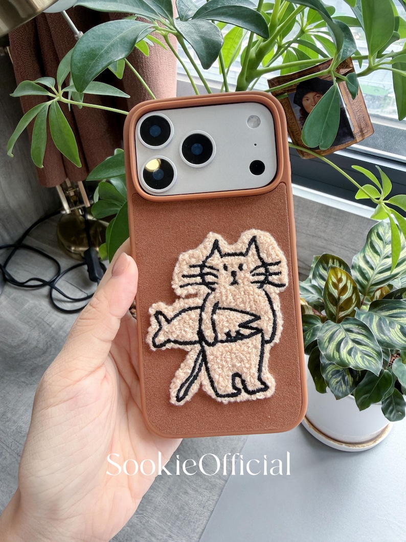 Puede incluir: Una funda de tel&eacute;fono marr&oacute;n con un dise&ntilde;o de gato beige texturizado. La funda tiene un recorte para la c&aacute;mara y el texto "SookieOfficial" en la parte inferior. La funda del tel&eacute;fono se sostiene en una mano, con plantas verdes en el fondo.