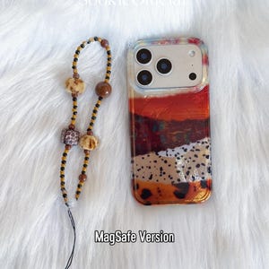 May include: A clear phone case with a colorful abstract design in shades of red, orange, brown, and beige. A beaded phone charm with brown, black, and beige beads is next to the phone case. The text "MagSafe Version" is at the bottom.