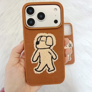 Puede incluir: Funda de tel&eacute;fono marr&oacute;n con un aplique de perro beige. La funda tiene un borde marr&oacute;n y una parte trasera texturizada. El aplique del perro est&aacute; delineado en negro. La marca "SookieOfficial" est&aacute; impresa en la parte superior.