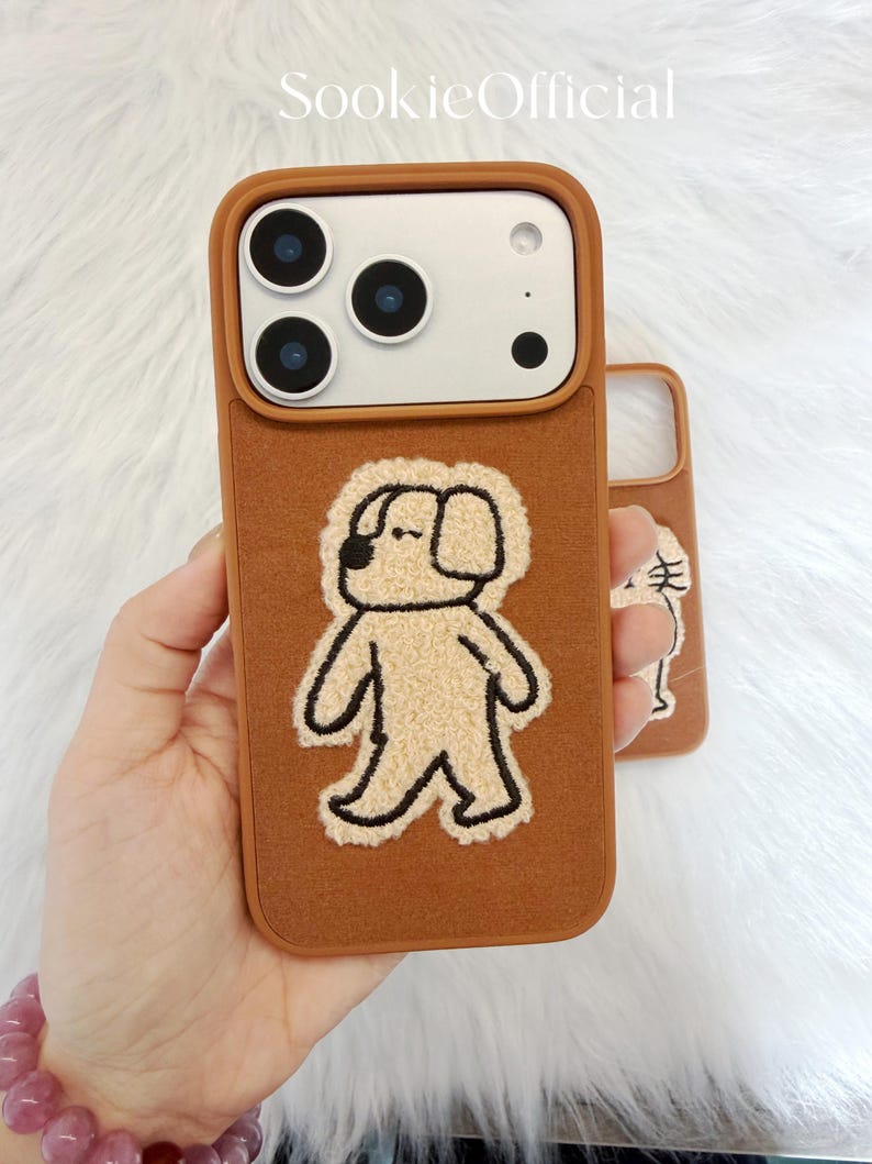 Puede incluir: Funda de tel&eacute;fono marr&oacute;n con un aplique de perro beige. La funda tiene un borde marr&oacute;n y una parte trasera texturizada. El aplique del perro est&aacute; delineado en negro. La marca "SookieOfficial" est&aacute; impresa en la parte superior.