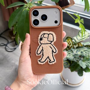 Puede incluir: Una funda de tel&eacute;fono marr&oacute;n con una aplicaci&oacute;n de perro beige y esponjoso. La funda tiene un borde marr&oacute;n y una parte trasera texturizada. La funda del tel&eacute;fono se sostiene en la mano de una persona, con una pulsera visible en primer plano. El texto "SookieOfficial" est&aacute; impreso en la funda.