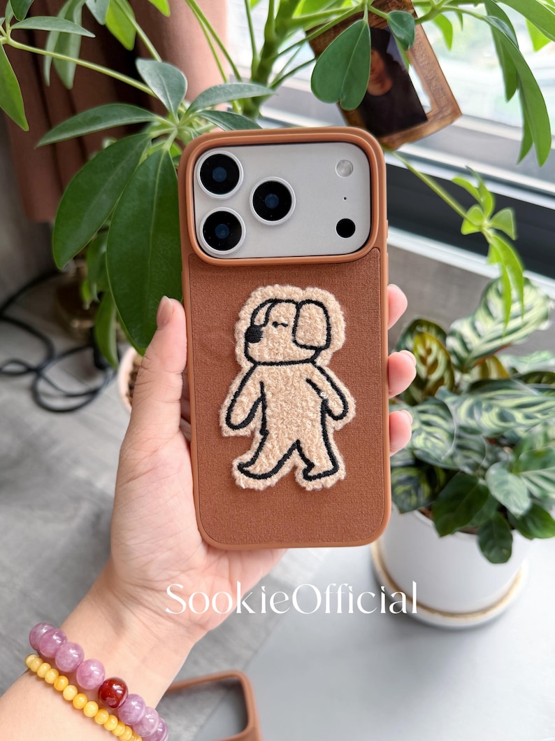 Puede incluir: Una funda de tel&eacute;fono marr&oacute;n con una aplicaci&oacute;n de perro beige y esponjoso. La funda tiene un borde marr&oacute;n y una parte trasera texturizada. La funda del tel&eacute;fono se sostiene en la mano de una persona, con una pulsera visible en primer plano. El texto "SookieOfficial" est&aacute; impreso en la funda.