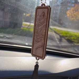 Puede incluir: Un amuleto de coche de madera con un borla negra colgando. El amuleto presenta un diseño de dragón tallado con láser.