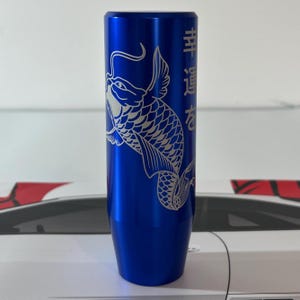 以下が含まれることがあります： 銀色の鯉のデザインと日本語の文字が入った青い円筒形のシフトノブ。金属製のシフトノブは、滑らかで反射する表面を持っています。鯉は鱗とひれで詳細に描かれています。