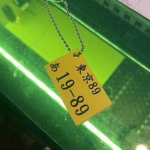 Könnte beinhalten: Ein gelbes Schild mit schwarzem Text, auf dem "89 19-89" steht, hängt an einer silbernen Kette.