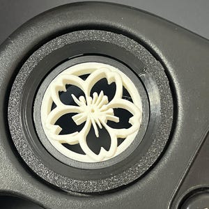 May include: A cream-coloured, flower-shaped object with detailed petals is set within a black, circular frame. The object is centred, and the background is a dark, neutral colour. The design is intricate and appears to be a decorative element.