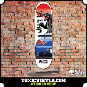 Puede incluir: Una tabla de skate blanca con un gráfico azul y negro de un coche y un monstruo parecido a Godzilla. El texto "ゴジラ" está impreso en la tabla. El texto "TOXIC VINYLS" está impreso en la parte inferior de la tabla.