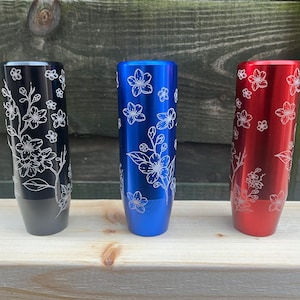 Pode incluir: Três manípulos de câmbio metálicos em preto, azul e vermelho. Cada manípulo é cilíndrico com um design floral branco gravado. Os manípulos são exibidos em uma superfície de madeira contra um fundo de madeira.